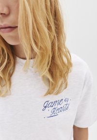 T-shirt blanche avec "jeu de cœurs" brodé en bleu. Texture douce, coupe décontractée, design minimal, décolleté et zone des épaules visibles.