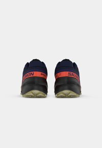 Sapatos de corrida em trilhos em azul-marinho e vermelho, com uma sola texturizada preta e um padrão de cravos proeminente. Apresentam um laço na parte de trás.
