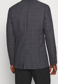 Tmavě kostkovaný blazer s texturovanou tkaninou, s větracími otvory na zadní straně a kontrastními knoflíky na manžetách, dobře padnoucí.