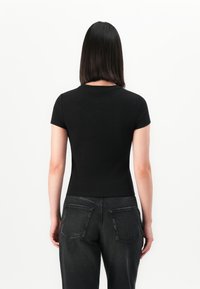 Woman with shoulder-length black hair wearing a fitted black short-sleeve top and dark high-waisted jeans, standing facing away.