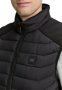 Chaleco acolchado negro con cuello alto, con segmentos acolchados horizontales y un pequeño parche con el logo en el lado izquierdo. Detalle texturizado en los hombros.