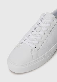 White leather sneaker with a rounded toe, flat rubber sole, and flat white laces. Features embossed branding on the side and stitching detail.