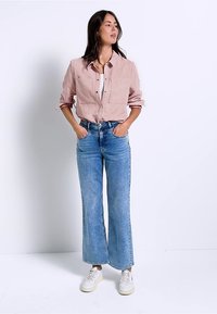 Světle růžová košile na knoflíky se dvěma náprsními kapsami, doplněná o světle modré rozšířené džíny a bílé tenisky. Model stojí se založenýma rukama v kapsách.