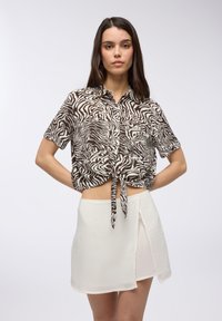 Camicetta a maniche corte a strisce zebra marrone e bianco legata in vita, abbinata a una gonna avvolgente bianca con uno spacco diagonale.