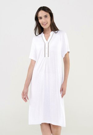 Mujer con cabello largo y oscuro, vistiendo un vestido blanco hasta la rodilla, con mangas cortas y detalles decorativos cerca del escote con botones, sonriendo.