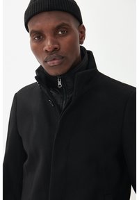 Cappotto nero con colletto alto e zip, caratterizzato da un design a strati con una sezione interna gonfiata. Tessuto liscio con dettagli hardware minimi.