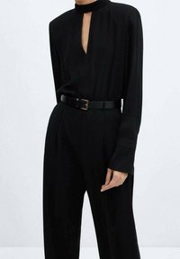 Combinaison noire avec un col haut, décolleté en forme de clé, manches longues, taille cintrée par une ceinture noire et pantalons larges. Texture lisse.