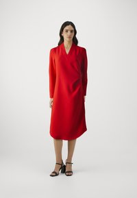 Rotes, langärmeliges Wickelkleid mit V-Ausschnitt und weichem Material, mit gerafften Schultern und lockerem Schnitt, kombiniert mit schwarzen, hochhackigen Sandalen.