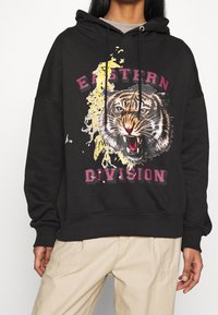 Svart huvtröja med en tiger grafik och texten "EASTERN DIVISION" i vinröd. Tillverkad i bomull med en avslappnad passform och dragsko.