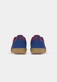 Blå og røde Nike sneakers med en overdel i ruskind, gummisål og tekstureret greb. "NIKE" logo i rødt på hælen. Rund form, lavt skåret design.