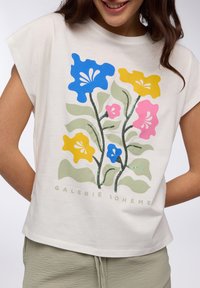 Maglietta bianca a maniche corte con una stampa floreale colorata in blu, giallo, rosa e verde, con il testo "GALERIE BOHÈME" in verde chiaro.