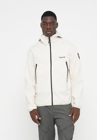 Lys beige zip-jakke med høj krave, to sidelommer med lynlås og sorte lynlåsdetaljer, parret med grå cargo-bukser.