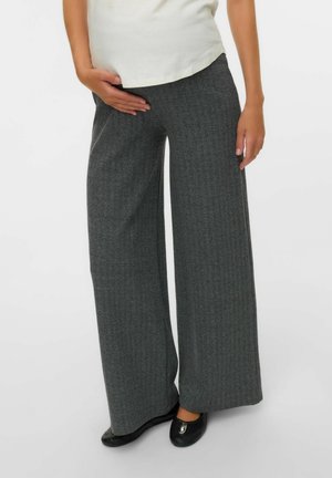 Person trägt lockere, graue Herringbone-Weitbein-Hose, weißes Oberteil und schwarze Ballettschuhe und steht vor weißem Hintergrund.