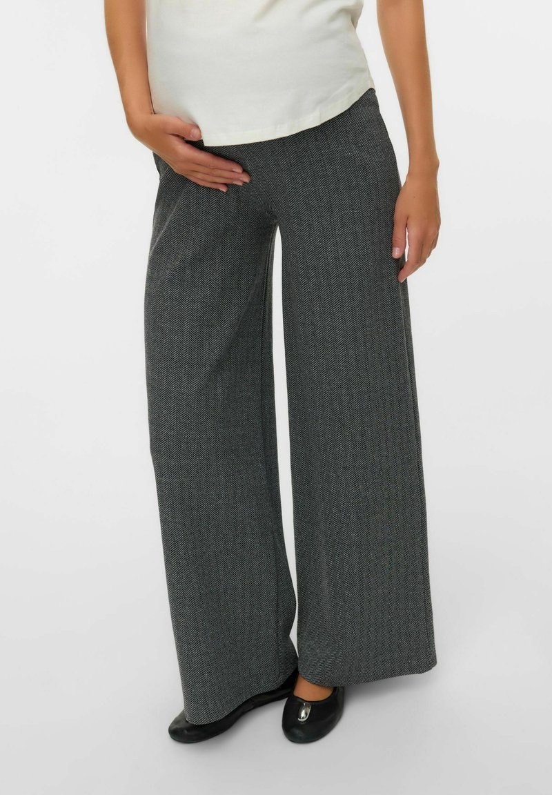 Personne portant un pantalon large ample en chevrons gris, un haut blanc et des ballerines noires, debout sur un fond blanc.