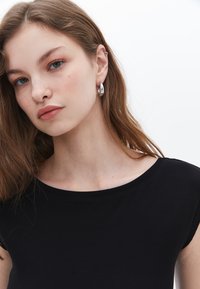Svart ribbad topp med en bred halsringning. Modell som bär silverfärgade hoops, visar långt vågigt hår och naturlig makeup mot en vit bakgrund.
