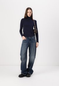 Marineblå høyhalset genser, slitte vidbukser i denim, svarte klumpete sko og en liten brun skulderveske. Minimalistisk design, moderne passform.