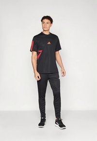 Conjunto de roupa desportiva preta composto por t-shirt com gola redonda, detalhes vermelhos e logótipo, combinada com calças de treino pretas ajustadas e ténis pretos.