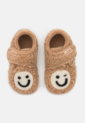 Chaussons d’enfant marron doux avec des sangles ajustables, chacun avec un patch blanc avec un visage souriant — l’un faisant un clin d’œil — sur les orteils.