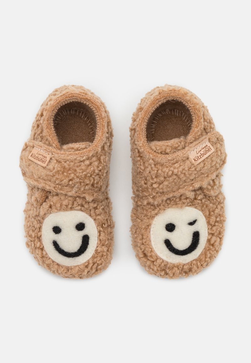Chaussons d’enfant marron doux avec des sangles ajustables, chacun avec un patch blanc avec un visage souriant — l’un faisant un clin d’œil — sur les orteils.
