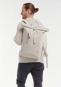 Rucksack mit Rollverschluss aus hellgrauem Material, verfügt über eine vordere Reißverschlusstasche mit farblichen Akzenten, verstellbareTräger und ein Logo an der Seite.