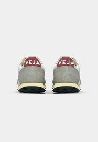 Sneaker bianche e grigie con tallone in camoscio, suola gialla e dettagli rossi. Logo "VEJA" sul retro, design minimalista.