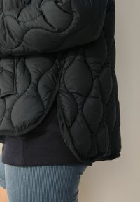 Giacca nera trapuntata con un design testurizzato e imbottito. Presenta un orlo a coste in contrasto ed è sovrapposta a pantaloni a coste blu chiaro.