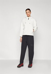 Ženska, ki stoji oblečena v belo flis jakno z kapuco, črne cargo hlače in rdeče pohodniške čevlje, proti enobarvni svetlo sivi ozadju.