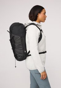 Svart ryggsäck med en elegant, böjd design, med justerbara remmar, dragkedjefickor och slitstarkt material. Bärs över en grå hoodie.