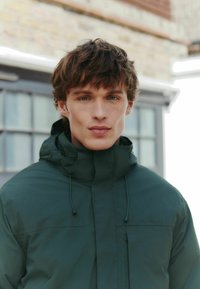 Dark green waterproof jacket with a high collar, adjustable drawstring, and smooth texture. Features a front zipper and pockets.