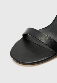 Gros plan sur l’avant d’une sandale en cuir noir avec une large bride sur les orteils sur un fond neutre.