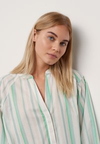 Blouse blanche avec des rayures vert clair et bleu, dotée d'un décolleté en V et de manches amples. Le tissu semble léger et texturé.
