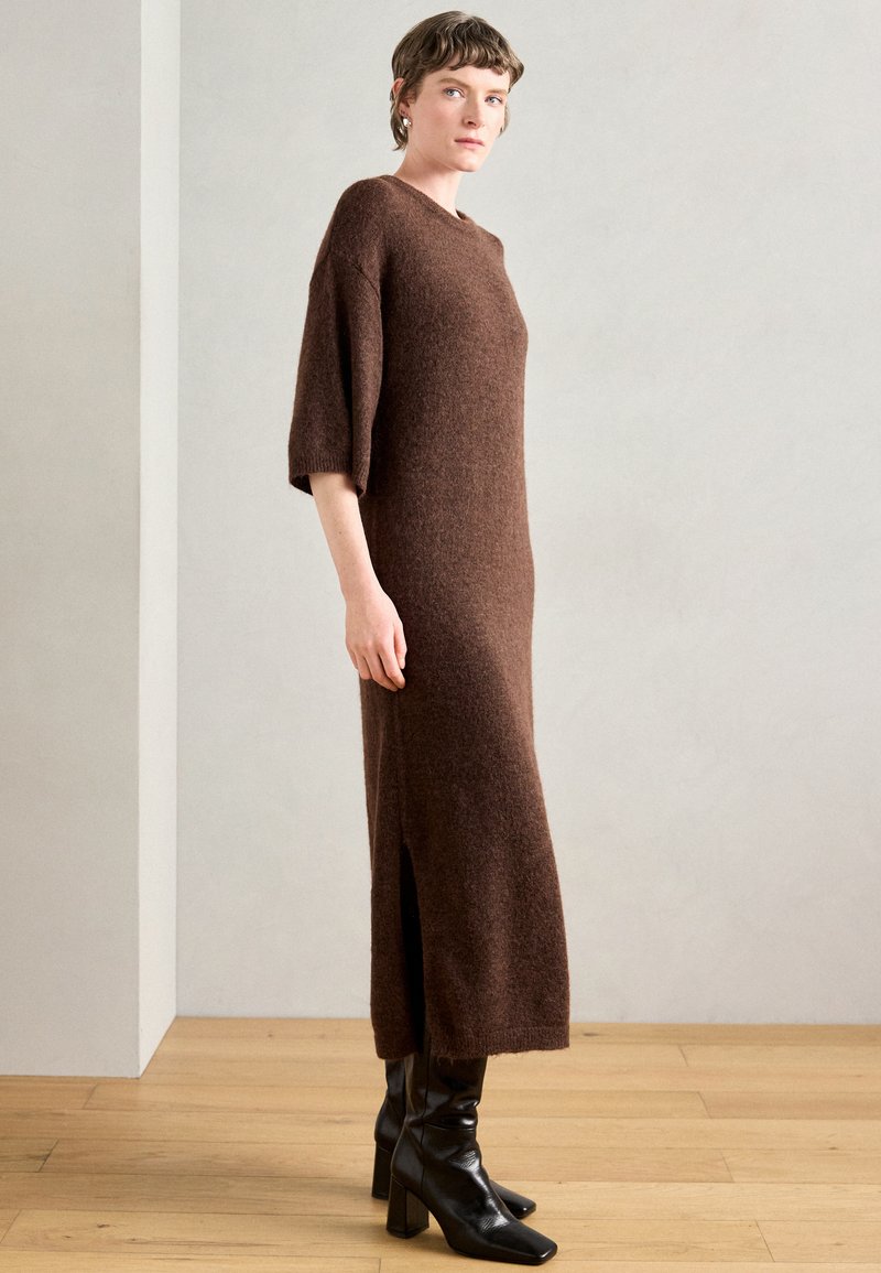 Brūns adīts kleitas tops ar apaļu dekoltē, trīs ceturtdaļu piedurknēm un sānu šķēlumu, savienots ar melnajām papēžu zābakiem. Strukturēta auduma.