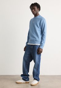 Ανοιχτό μπλε φούτερ με ribbed μανσέτες και σακάκι, φορεμένο πάνω από ένα άσπρο �πουκάμισο. Συνδυασμένο με φαρδιά μπλε τζιν και λευκά αθλητικά παπούτσια με μπεζ σόλες.