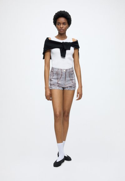 Femme portant un haut blanc sans manches, un short taille haute à motifs, un pull noir noué autour des épaules, des chaussettes blanches et des ballerines noires, debout sur un fond blanc.