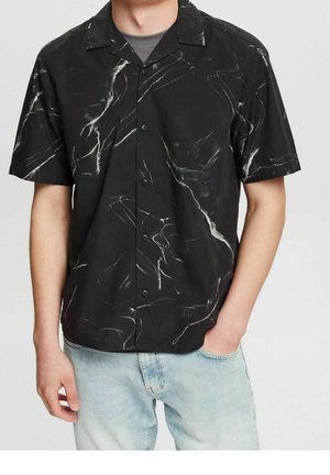 Homme portant une chemise noire à manches courtes avec un motif blanc marbré et un jean bleu clair, debout avec une main dans la poche.