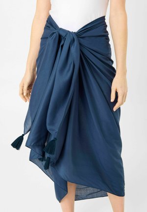 Frau trägt einen marineblauen Sarong, der mit einer Schleife an der Taille mit Quasten gebunden ist, kombiniert mit einem weißen ärmellosen Top.