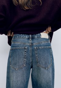 Högmidjade blå denimjeans med en urtvättad finish, försedda med en vit märkestagg på midjan och två bakfickor.