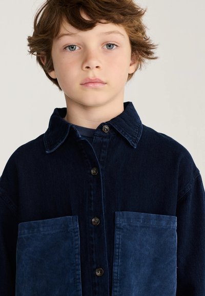 Garçon aux cheveux châtain clair et aux yeux bleus, portant une chemise bleu foncé boutonnée avec deux grandes poches à l'avant, sur un fond clair uni.