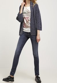 Mörkblå stickad cardigan, vit grafiktröja och mörka smala jeans. Svarta lackloafers med spänne. Enkel, avslappnad outfit.