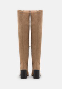 Botine din suede bej, până la genunchi, cu o textură netedă, design drept și toc bloc negru. Au fermoare laterale pentru închidere.