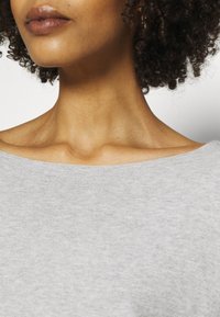 Close-up van het ondergezicht en de nek van een persoon met krullend haar, die een lichtgrijze off-shoulder top draagt.