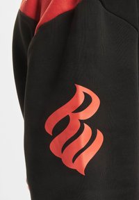 Svart hettegenser med røde detaljer; har et stilisert flamme-design i rødt på ermene. Glatt tekstur, avslappet passform.
