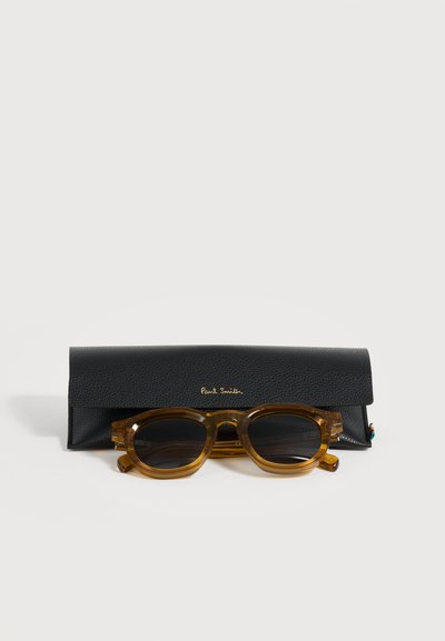 Lunettes de soleil translucides marron avec verres foncés placées devant un étui noir texturé de marque Paul Smith sur une surface blanche.