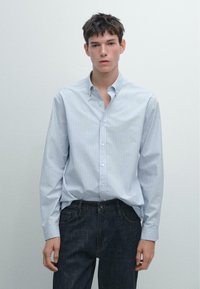 Camicia azzurra chiara con sottili righe verticali bianche, maniche lunghe e vestibilità rilassata, abbinata a jeans di denim scuro.