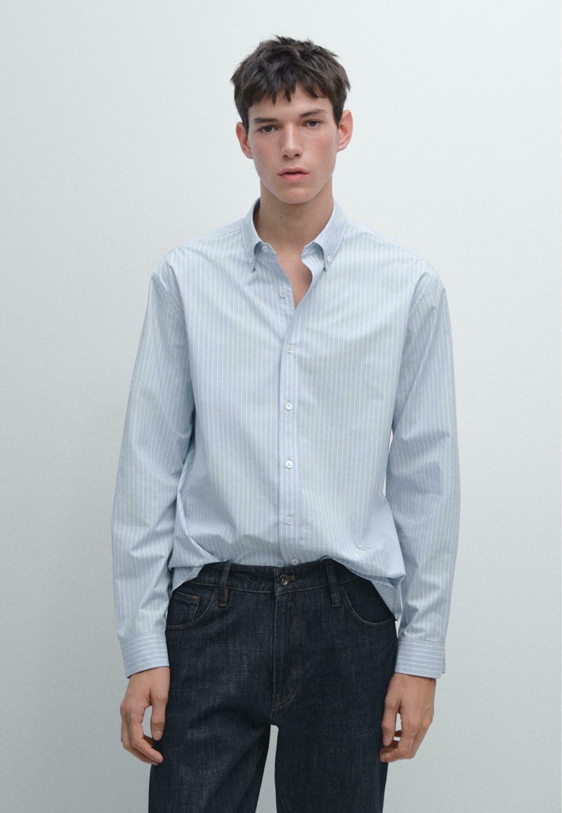 Camicia azzurra chiara con sottili righe verticali bianche, maniche lunghe e vestibilità rilassata, abbinata a jeans di denim scuro.