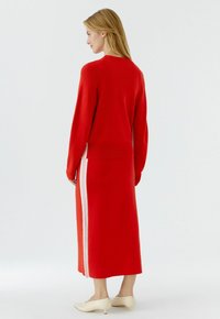 Frau trägt ein rotes Langarmoberteil und einen passenden Rock mit weißen Seitenstreifen, steht in cremefarbenen, flachen Schuhen vor weißem Hintergrund.