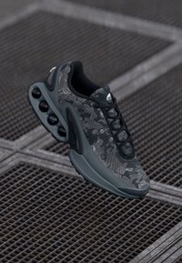 Mustat ja harmaat tennarit, joissa on teksturoitu maastokuvio, pyöreät ilmatyynybotit ja virtaviivainen muotoilu, jossa on nauhakiinnitys edessä.