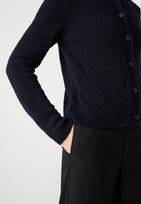 Femme portant un cardigan noir texturé à boutons avec une poche avant, main dans la poche d'un pantalon noir, sur un fond clair uni.