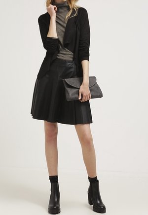 Woman wearing black cardigan, gray turtleneck, black leather skirt, black ankle boots, holding gray clutch purse, standing against white background.