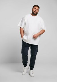 Vit bomull T-shirt med korta ärmar matchad med marinblå cargobyxor med fickor. Modellen bär vita sneakers och står mot en grå bakgrund.